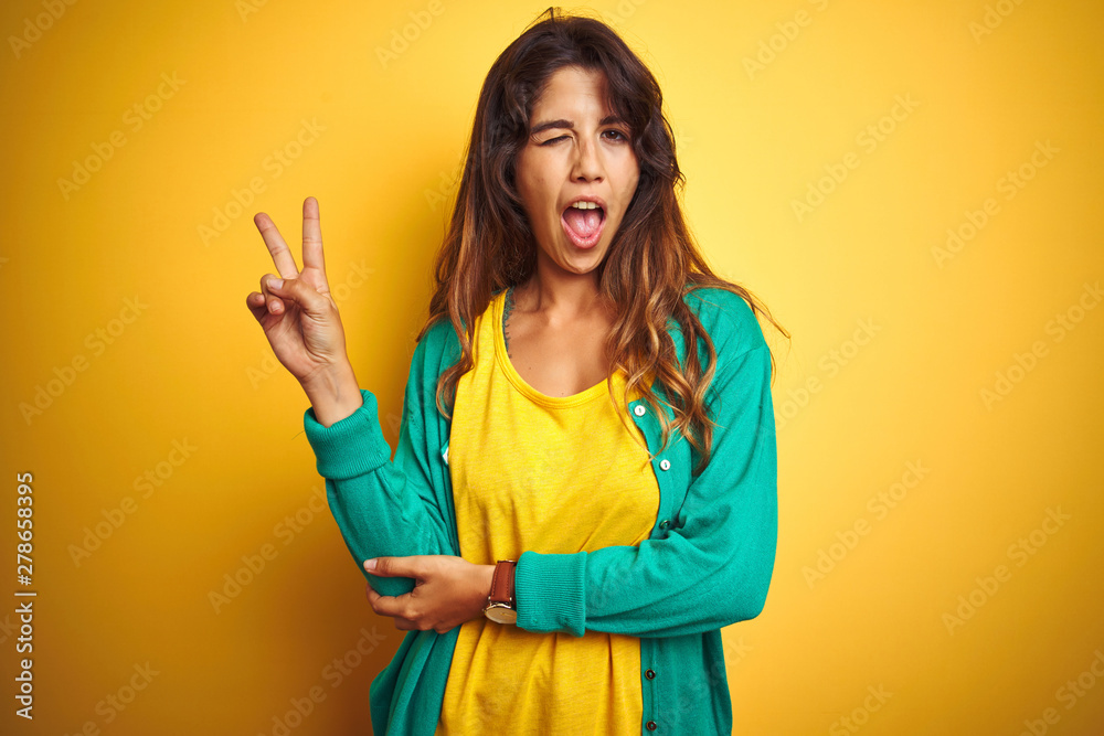 Young woman wearing t-shirt and green sweater standing over yelllow isolated background smiling with happy face winking at the camera doing victory sign. Number two. Young woman wearing t-shirt and green sweater standing over yelllow isolated background smiling with happy face winking at the camera doing victory sign. Number two.