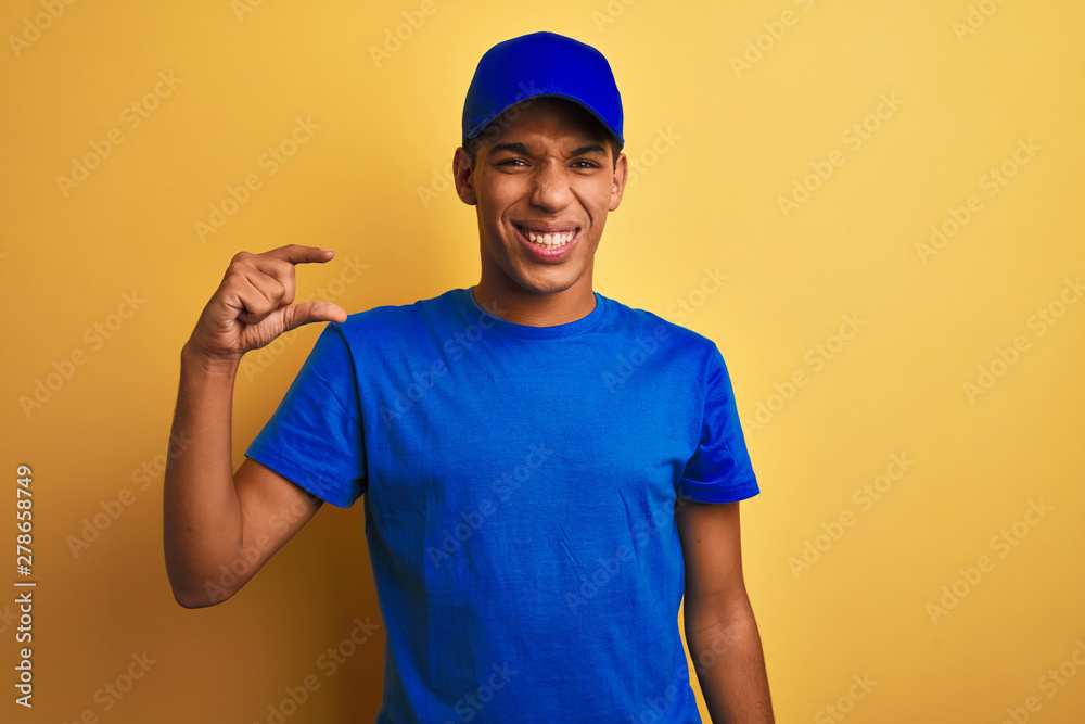 Young handsome arab delivery man standing over isolated yellow background smiling and confident gesturing with hand doing small size sign with fingers looking and the camera. Measure concept.