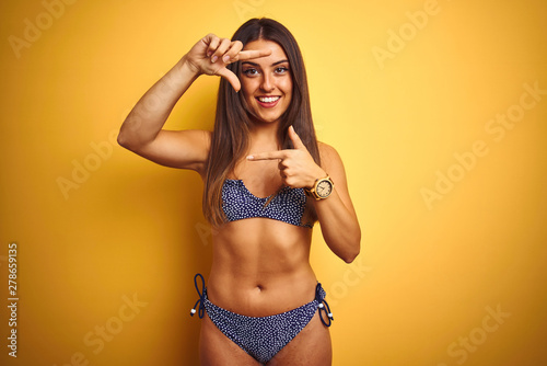 Young beautiful woman on vacation wearing bikini standing over isolated yellow background smiling making frame with hands and fingers with happy face. Creativity and photography concept.
