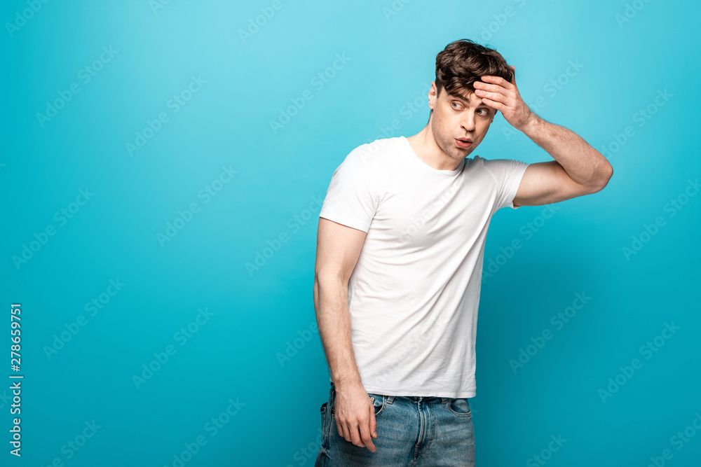 shocked young man looking away while holding hand on head on blue background