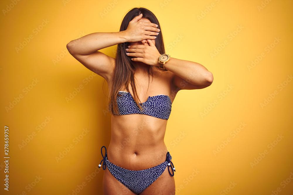 Naklejka premium Young beautiful woman on vacation wearing bikini standing over isolated yellow background Covering eyes and mouth with hands, surprised and shocked. Hiding emotion