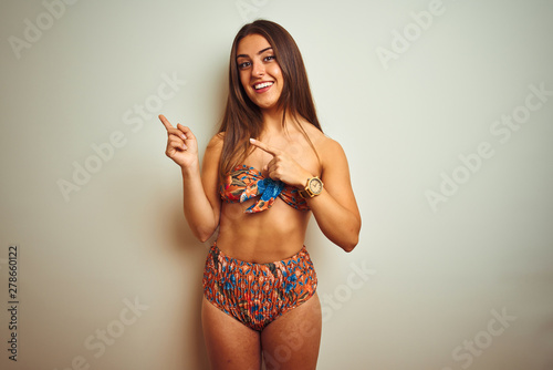 Young beautiful woman on vacation wearing bikini standing over isolated white background smiling and looking at the camera pointing with two hands and fingers to the side.