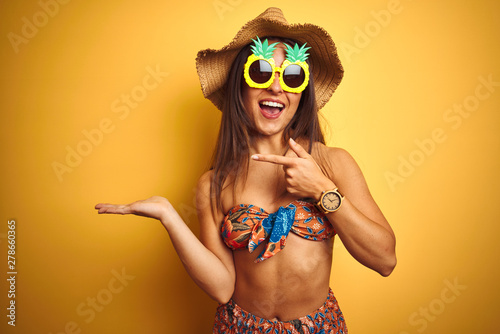 Woman on vacation wearing bikini and pineapple sunglasses over isolated yellow background amazed and smiling to the camera while presenting with hand and pointing with finger.