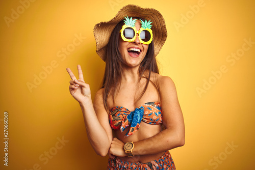 Woman on vacation wearing bikini and pineapple sunglasses over isolated yellow background smiling with happy face winking at the camera doing victory sign. Number two.