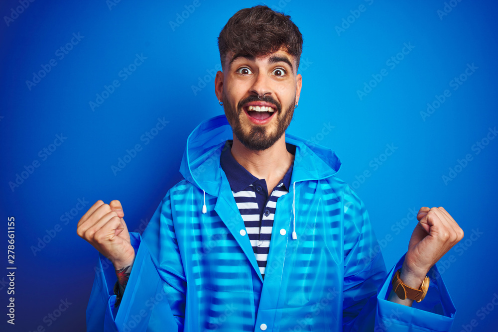 Young handsome man wearing rain coat standing over isolated blue background celebrating surprised and amazed for success with arms raised and open eyes. Winner concept.