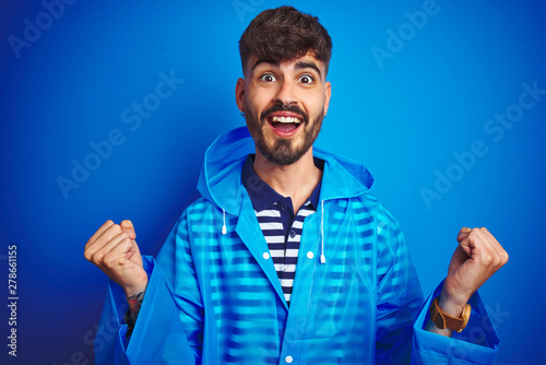 Young handsome man wearing rain coat standing over isolated blue background celebrating surprised and amazed for success with arms raised and open eyes. Winner concept.