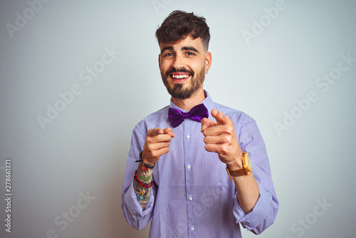 Young man with tattoo wearing purple shirt and bow tie over isolated white background pointing fingers to camera with happy and funny face. Good energy and vibes.