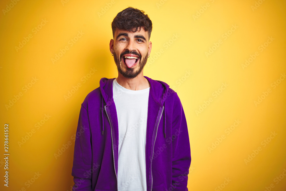 Young man with tattoo wearing sport purple sweatshirt over isolated yellow background sticking tongue out happy with funny expression. Emotion concept. - obrazy, fototapety, plakaty 