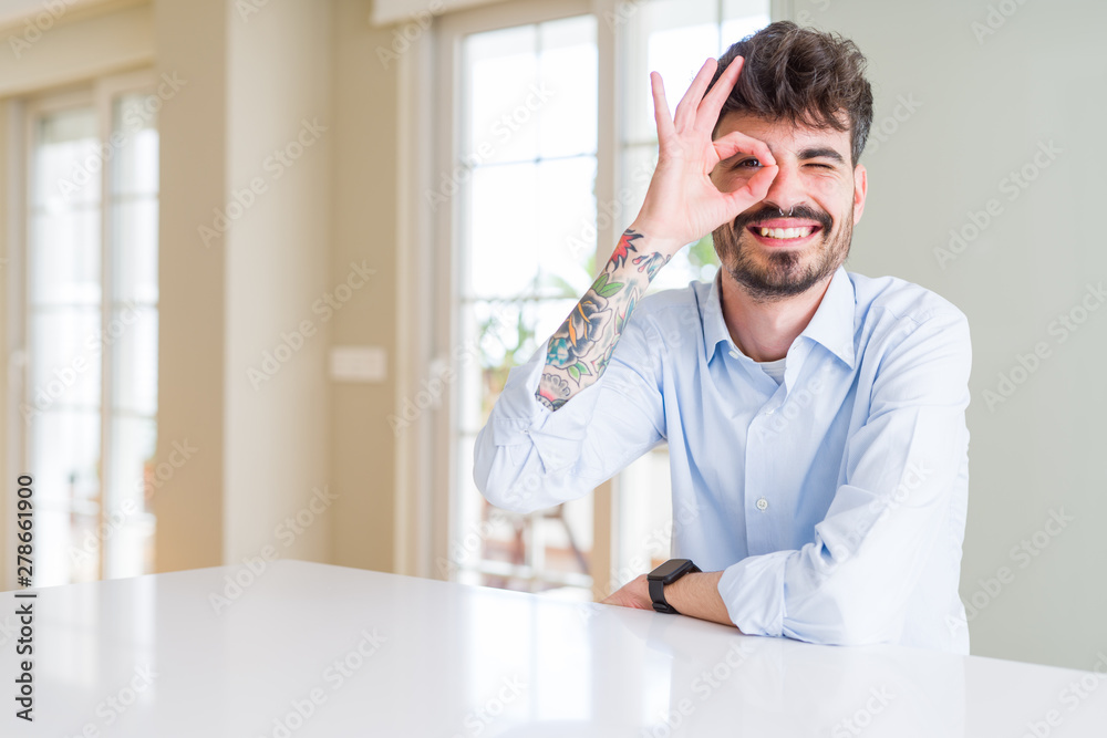 Young businesss man sitting on white table doing ok gesture with hand smiling, eye looking through fingers with happy face.