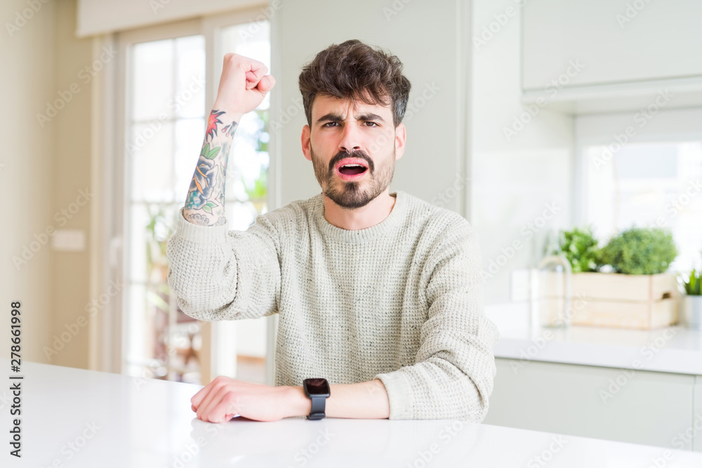 Young man wearing casual sweater sitting on white table angry and mad raising fist frustrated and furious while shouting with anger. Rage and aggressive concept.