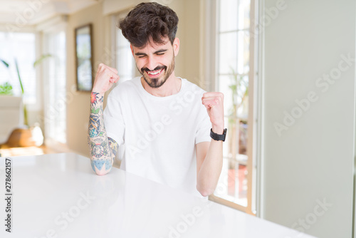 Young man wearing casual t-shirt sitting on white table very happy and excited doing winner gesture with arms raised, smiling and screaming for success. Celebration concept.