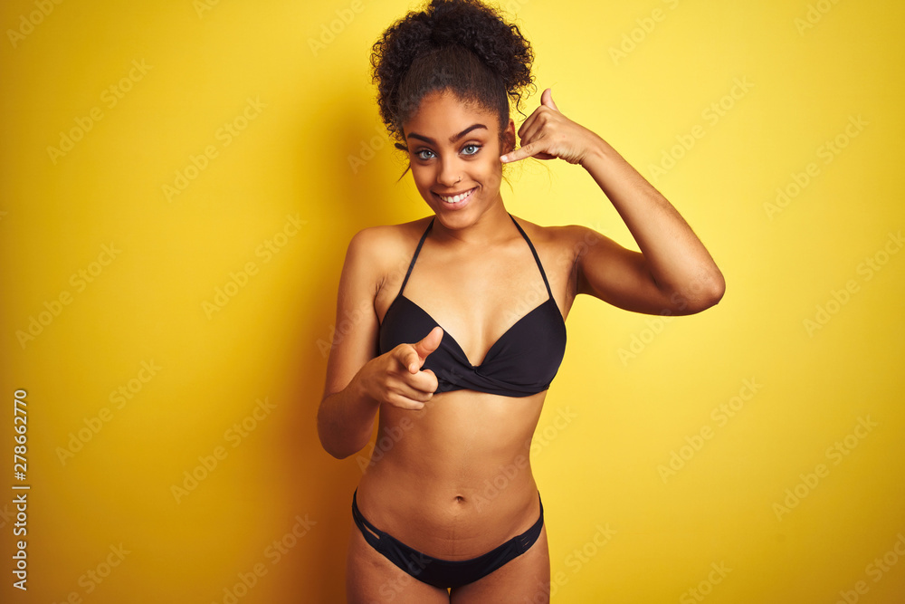 African american woman on vacation wearing bikini standing over isolated yellow background smiling doing talking on the telephone gesture and pointing to you. Call me. African american woman on vacation wearing bikini standing over isolated yellow background smiling doing talking on the telephone gesture and pointing to you. Call me.