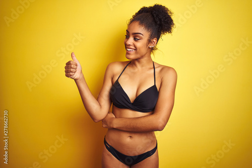 African american woman on vacation wearing bikini standing over isolated yellow background Looking proud, smiling doing thumbs up gesture to the side © Krakenimages.com