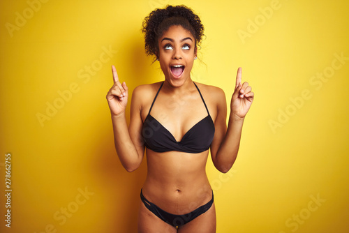 African american woman on vacation wearing bikini standing over isolated yellow background smiling amazed and surprised and pointing up with fingers and raised arms. © Krakenimages.com