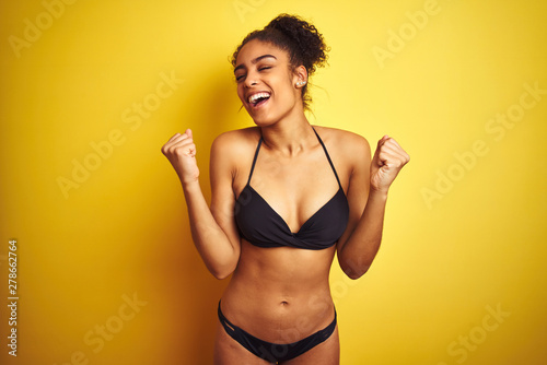 African american woman on vacation wearing bikini standing over isolated yellow background celebrating surprised and amazed for success with arms raised and eyes closed. Winner concept.