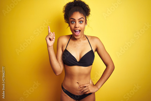 African american woman on vacation wearing bikini standing over isolated yellow background pointing finger up with successful idea. Exited and happy. Number one.