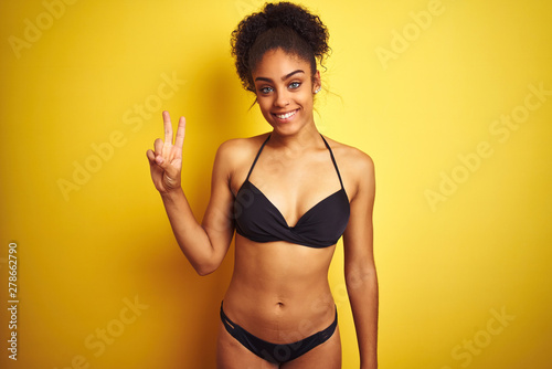 African american woman on vacation wearing bikini standing over isolated yellow background showing and pointing up with fingers number two while smiling confident and happy. © Krakenimages.com