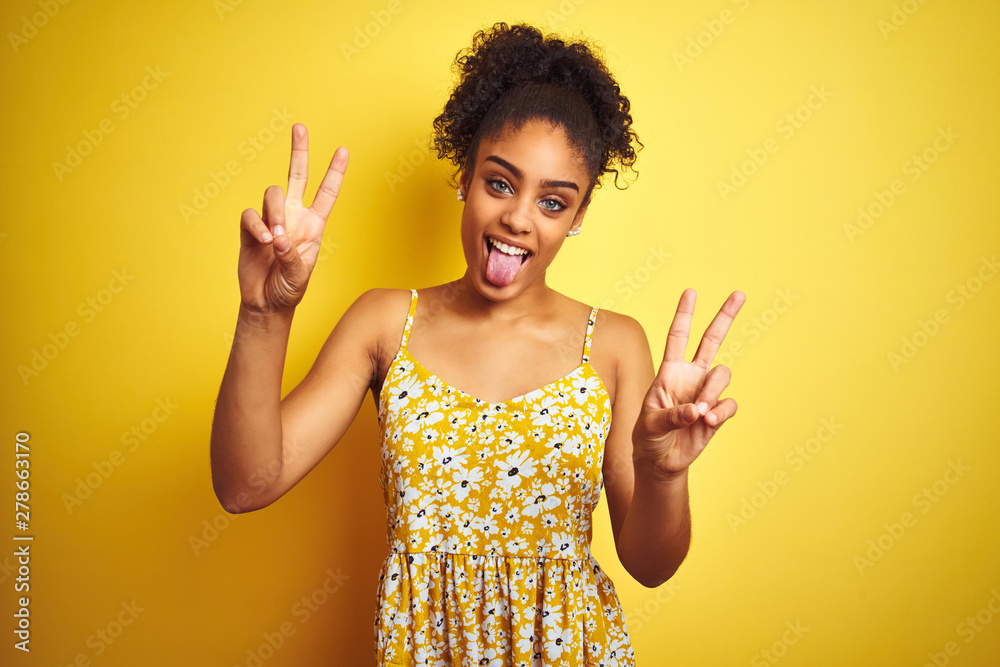 African american woman wearing casual floral dress standing over isolated yellow background smiling with tongue out showing fingers of both hands doing victory sign. Number two. African american woman wearing casual floral dress standing over isolated yellow background smiling with tongue out showing fingers of both hands doing victory sign. Number two.