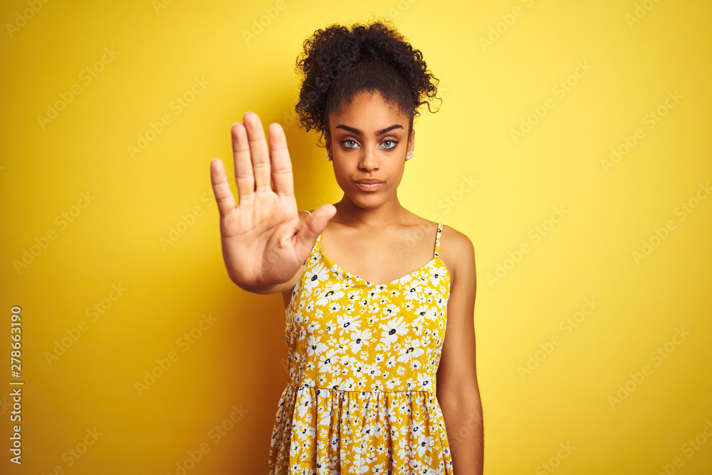 African american woman wearing casual floral dress standing over isolated yellow background doing stop sing with palm of the hand. Warning expression with negative and serious gesture on the face.