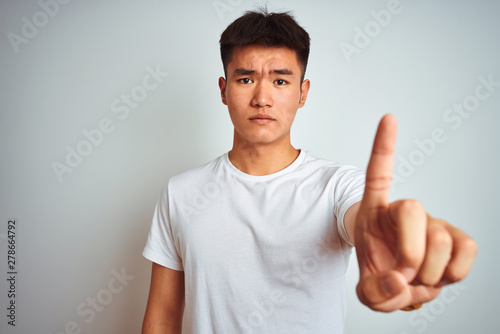 Young asian chinese man wearing t-shirt standing over isolated white background Pointing with finger up and angry expression, showing no gesture