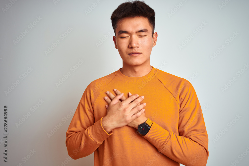 Young asian chinese man wearing orange sweater standing over isolated white background smiling with hands on chest with closed eyes and grateful gesture on face. Health concept.
