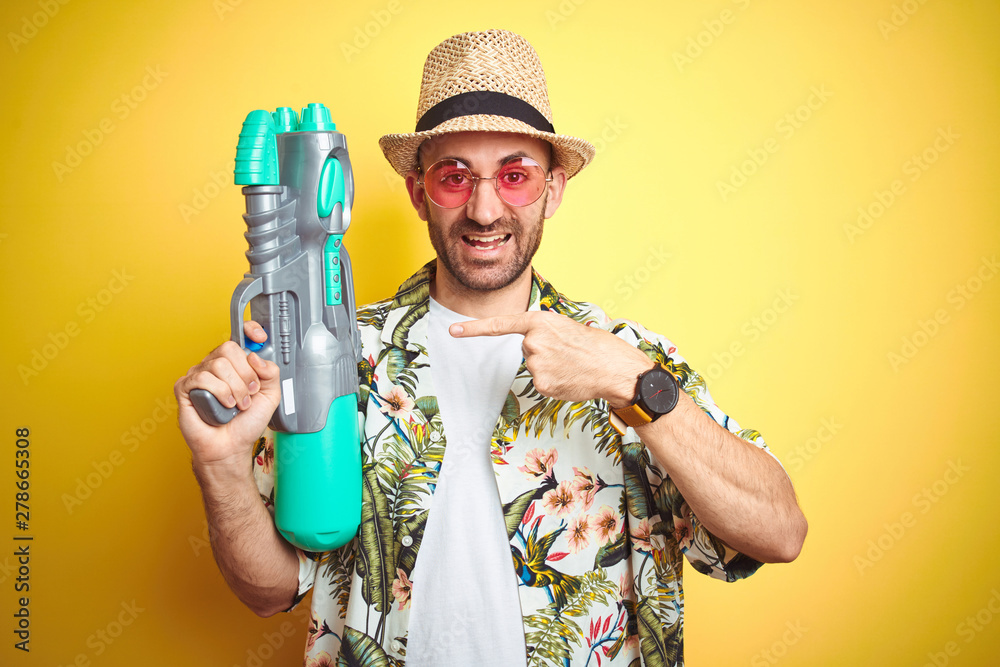 Young man wearing hawaiian flowers shirt holding water gun over yellow isolated background very happy pointing with hand and finger Young man wearing hawaiian flowers shirt holding water gun over yellow isolated background very happy pointing with hand and finger