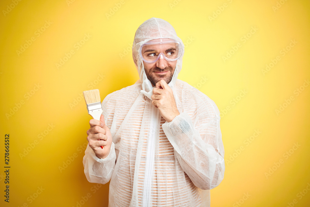 Young man wearing painter equipment and holding painting brush over isolated yellow background serious face thinking about question, very confused idea Young man wearing painter equipment and holding painting brush over isolated yellow background serious face thinking about question, very confused idea