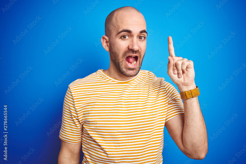Fototapeta premium Young bald man with beard wearing casual t-shirt over blue isolated background pointing finger up with successful idea. Exited and happy. Number one.