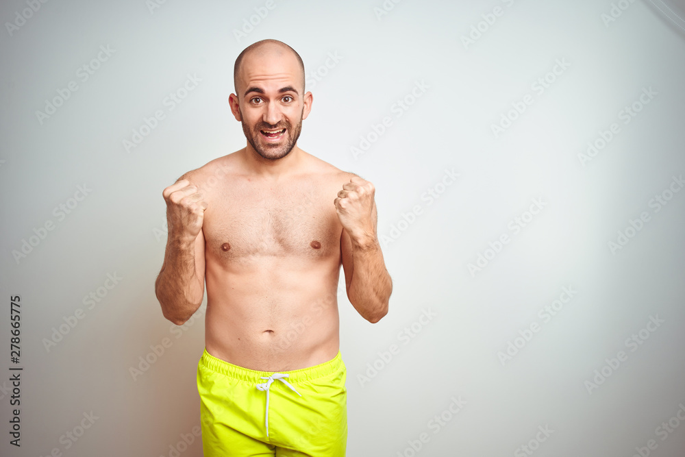 Young shirtless man on vacation wearing yellow swimwear over isolated background celebrating surprised and amazed for success with arms raised and open eyes. Winner concept. Young shirtless man on vacation wearing yellow swimwear over isolated background celebrating surprised and amazed for success with arms raised and open eyes. Winner concept.
