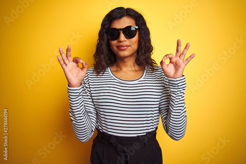 Transsexual transgender woman wearing sunglasses over isolated yellow background relax and smiling with eyes closed doing meditation gesture with fingers. Yoga concept.