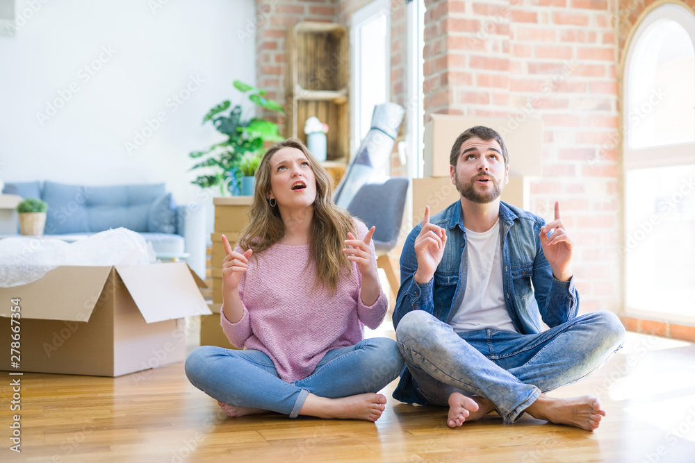Young beautiful couple moving to a new house sitting on the floor amazed and surprised looking up and pointing with fingers and raised arms.