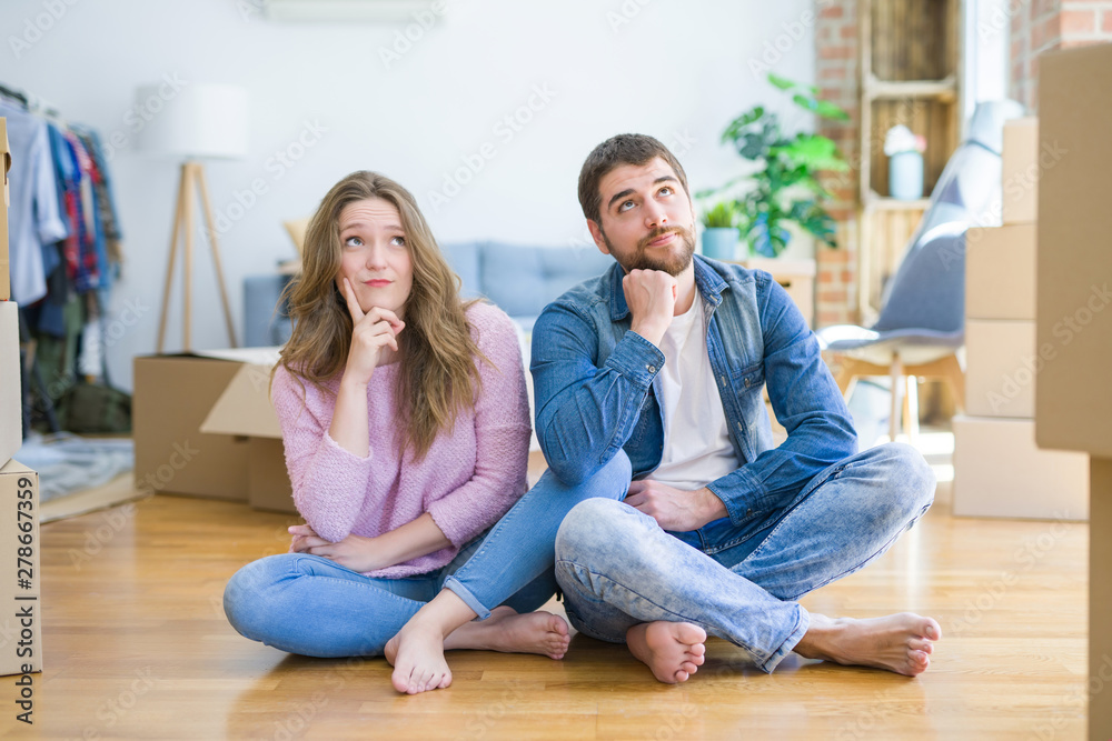 Young beautiful couple moving to a new house sitting on the floor with hand on chin thinking about question, pensive expression. Smiling with thoughtful face. Doubt concept.