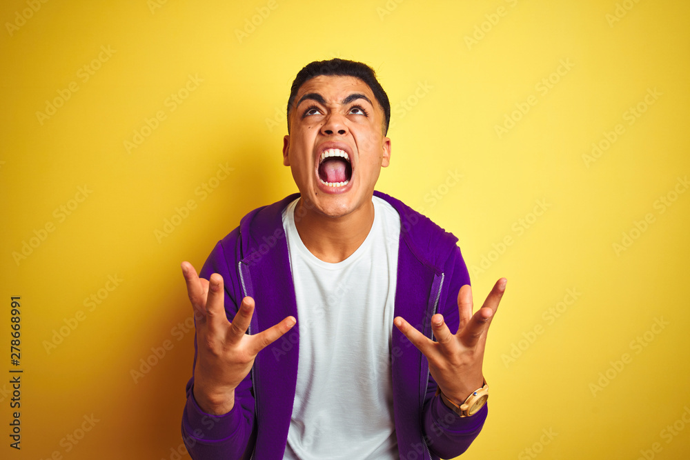 Fototapeta premium Young brazilian man wearing purple sweatshirt standing over isolated yellow background crazy and mad shouting and yelling with aggressive expression and arms raised. Frustration concept.