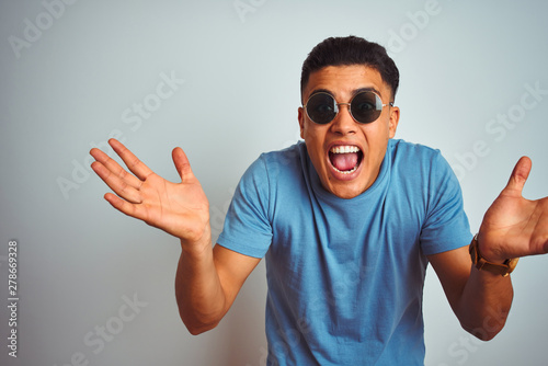 Young brazilian man wearing blue t-shirt and sunglasses over isolated white background celebrating crazy and amazed for success with arms raised and open eyes screaming excited. Winner concept