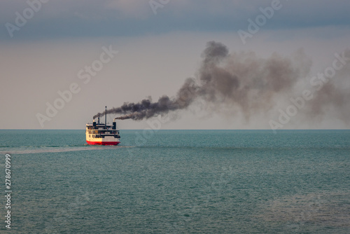 Huge boat or ferry with dark smoke float on the ocean at sunset time