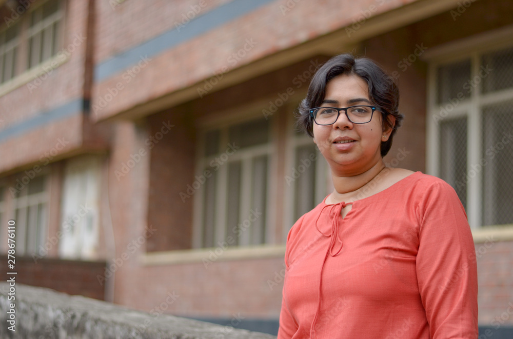 Fototapeta premium Portrait of a confident looking young Indian woman with short hair and spectacles, crossed folded hands in an outdoor setting wearing a traditional North Indian suit dress against a red brick wall
