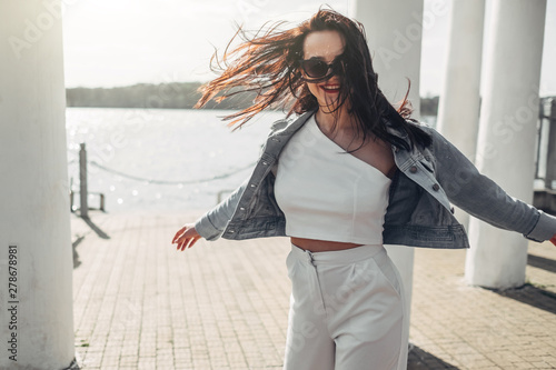 Beautiful Brunette Girl in Sunglasses White Pants and Jeans Jacket Near the Lake