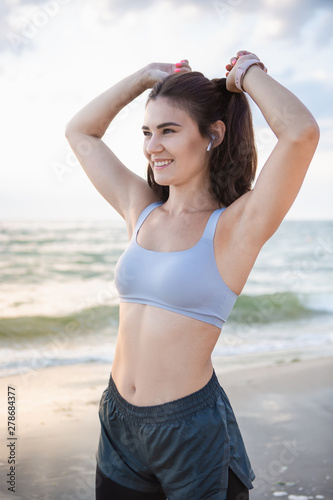Happy brunette woman fixes her hair after workout at the sea shore at sunrise. Model listening to the music during exercising near the sea.