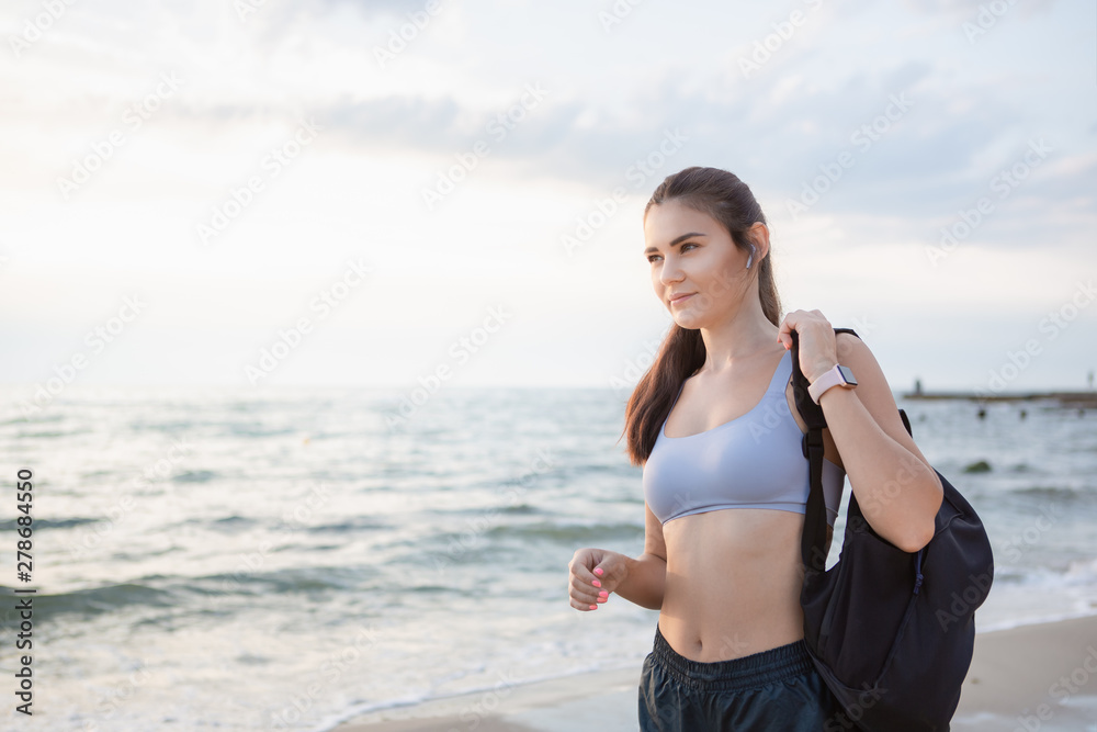 Young brunette woman with wireless earbuds, smart watches and black backpack resting after morning workout at the sea shore at sunrise. Young brunette woman with wireless earbuds, smart watches and black backpack resting after morning workout at the sea shore at sunrise.