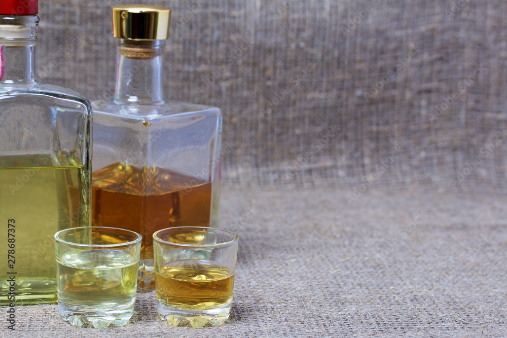 Bottled bottles of strong alcohol. From transparent glass, a square form. Near the stack with a drink poured. Against the backdrop of coarse linen fabric. Bottled bottles of strong alcohol. From transparent glass, a square form. Near the stack with a drink poured. Against the backdrop of coarse linen fabric.