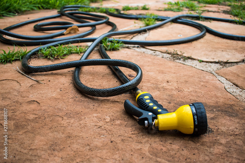 A black rubber hose with a brigh yellow nozzle randomly coiled across terracotta outdoor tiles and green grass.