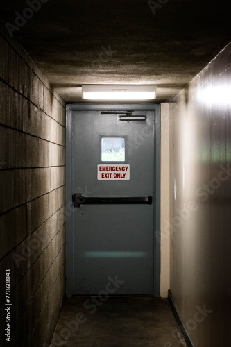 Emergency exit only sign on grey door at the end of a shadowed hallway with a single flourescent light above it.