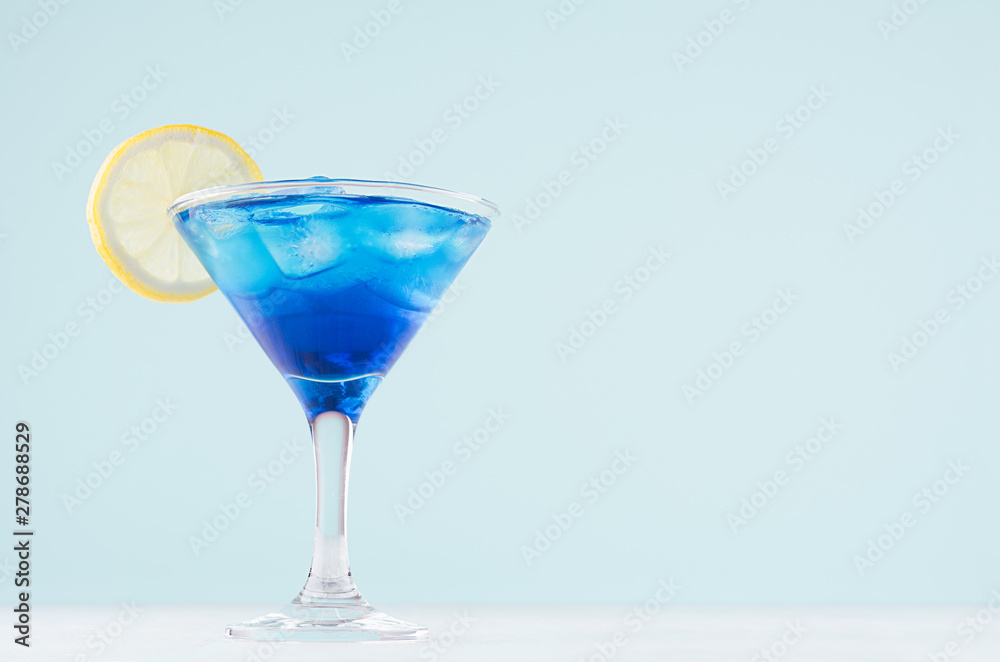 Tropical fresh alcohol cocktail with blue curacao liqueur, ice cube, lemon slice in misted martini glass on soft light white, pastel green background.