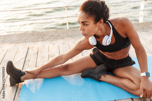Image of african american woman stretching her body on finess mat while doing workout by seaside in morning © Drobot Dean