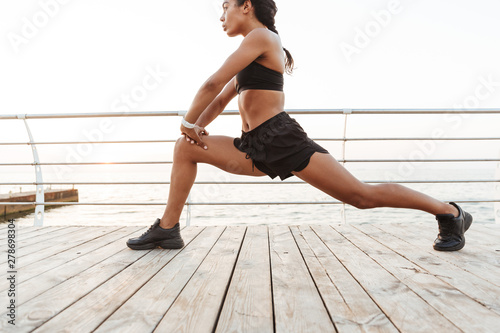 Image of focused young woman squatting and stretching her body while doing workout by seaside in morning