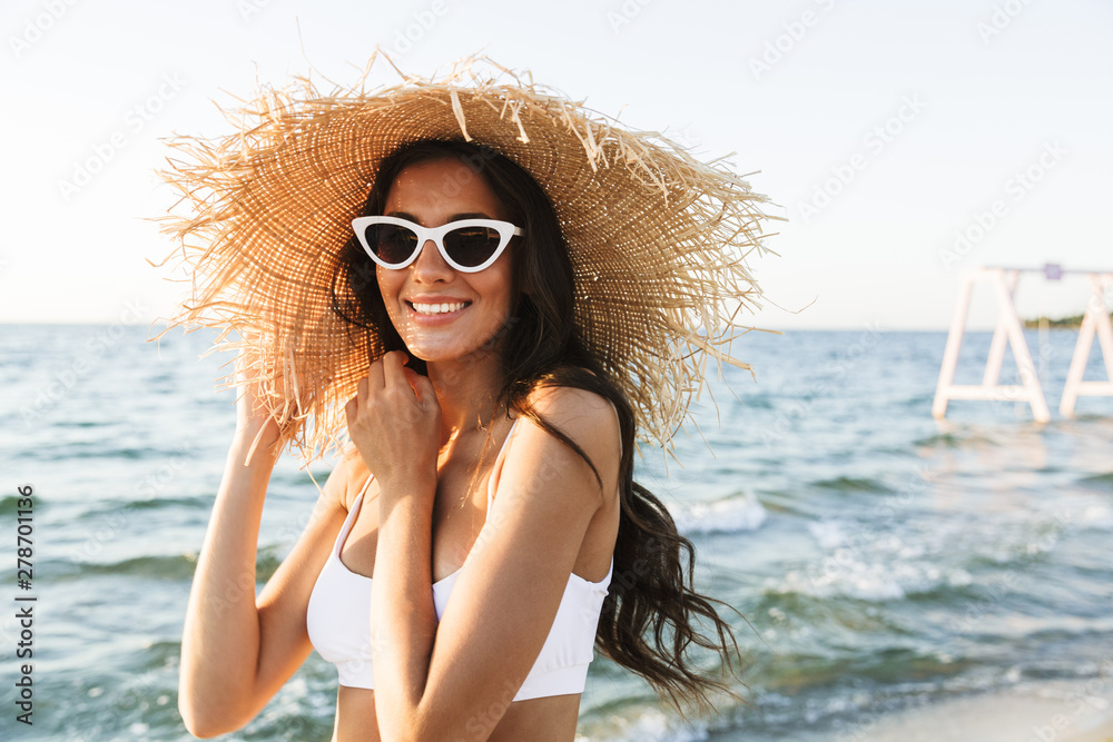 Photo of joyful brunette woman in swimsuit and straw hat smiling while walking by seaside Photo of joyful brunette woman in swimsuit and straw hat smiling while walking by seaside