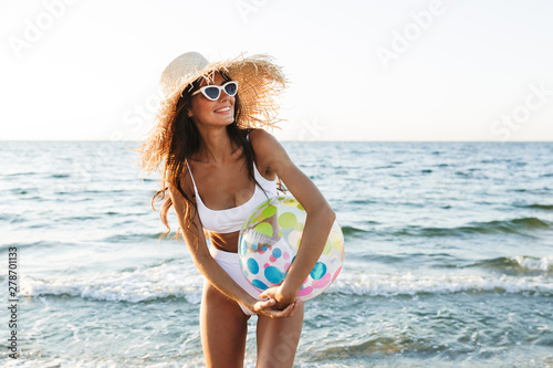 Fotografi Photo of cute brunette woman in swimsuit and straw hat holding beach ball while