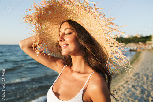 Photo of positive brunette woman in swimsuit and straw hat smiling and sunbathing while walking by seaside