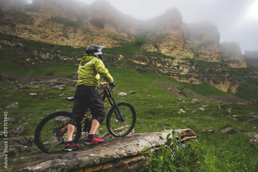 Fototapeta premium Side view of a man on a mountain bike standing on a rocky terrain and looking at a rock. The concept of a mountain bike and mtb downhill