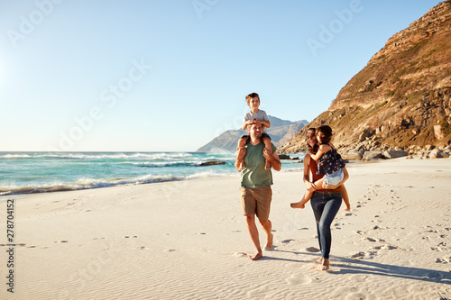 Mid adult white couple walking along on a beach on a family holiday carrying their kids, full length photo